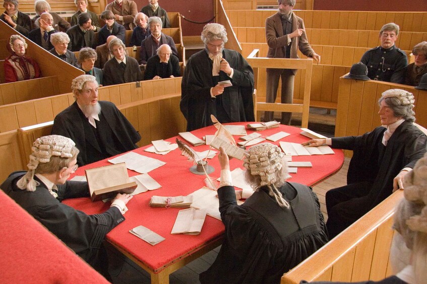 A courtroom with judges and a lawyer at a table, surrounded by seated audience. 3
