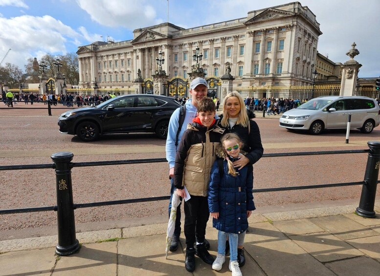 En familj poserar framför Buckingham Palace, med en svart bil och en vit skåpbil i bakgrunden. 3