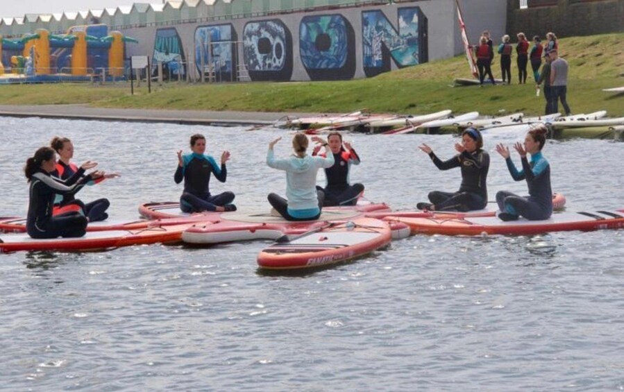 People sitting on paddleboards in a body of water, with a mural and a group of people in the background. 1