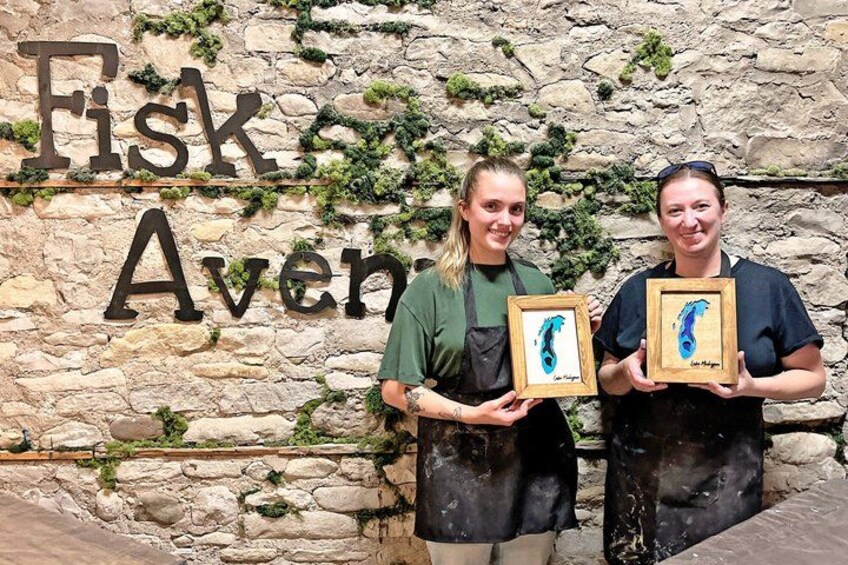 Two people holding framed artwork in front of a stone wall with the text 'Fisk Avenue'. 1