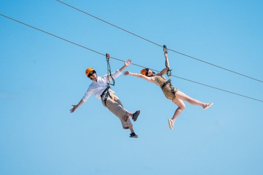 Two people ziplining, wearing helmets and harnesses, against a clear blue sky. 4