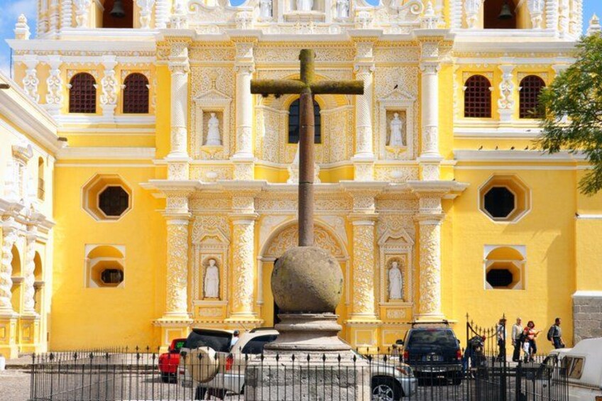A yellow colonial-style building with ornate architecture, a large stone cross, and a fence in the foreground. 4