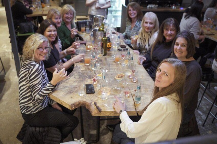 A group of people sitting around a wooden table with glasses and bottles, suggesting a social gathering or event. 3