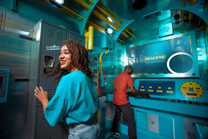 A person in a blue shirt and jeans standing in front of a control panel with the logo 'DESCEND' on it. 3
