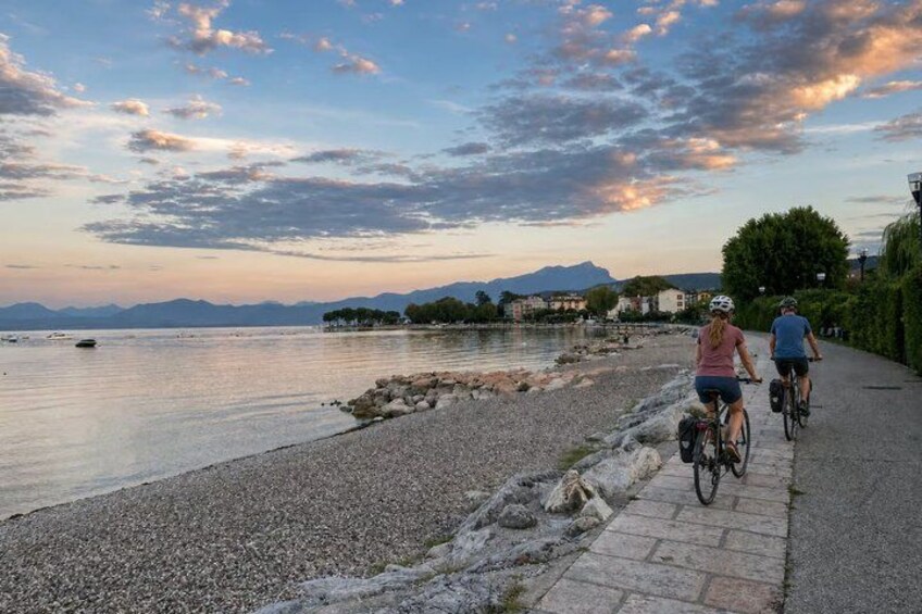 A river with boats, a pebbled beach, and cyclists on a path. 1