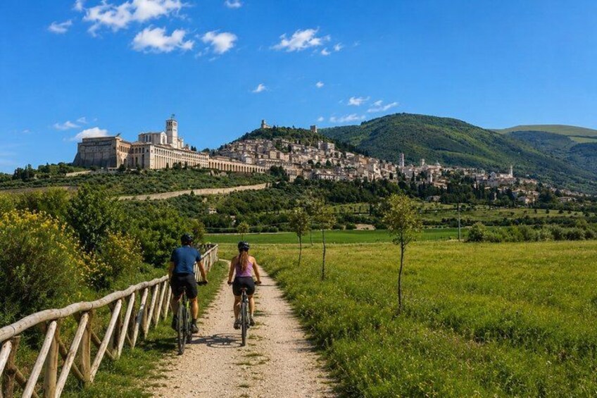 A gravel path with cyclists, a wooden fence, green fields, and a distant town with a prominent building. 1