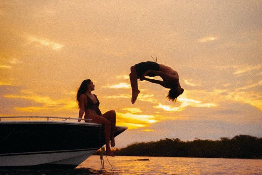 A sunset over a body of water with a boat and two people, one sitting and one diving. 5