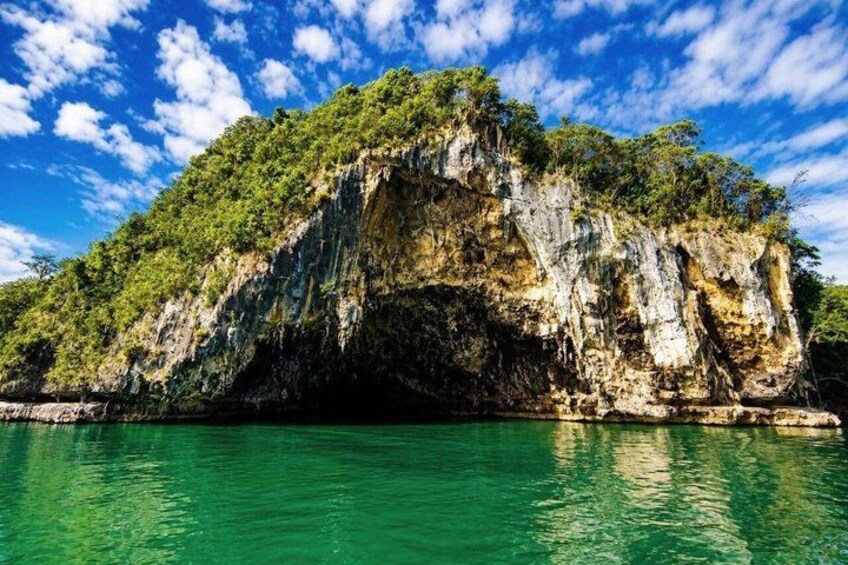 A coastal cliff with a cave entrance, surrounded by clear turquoise water and lush greenery. 1