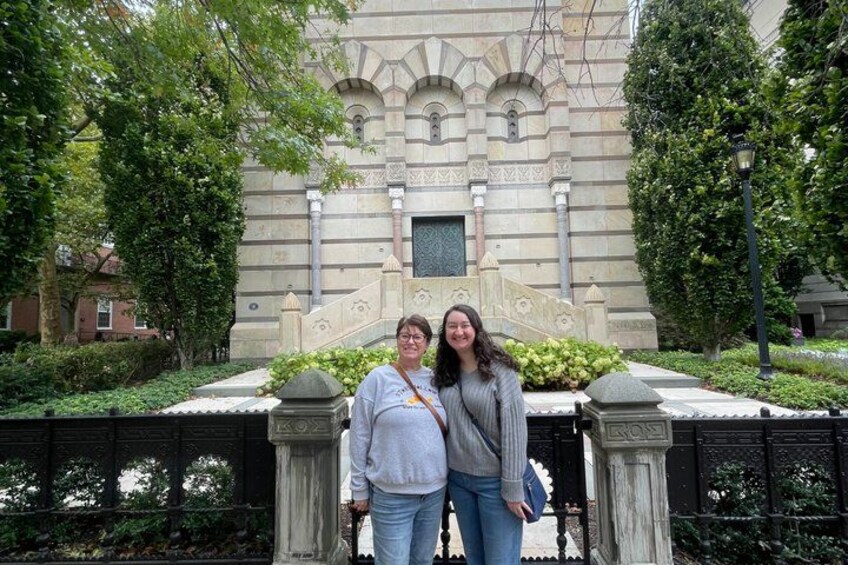 Two people standing in front of a building with ornate architecture, a stone gate, and greenery. 1