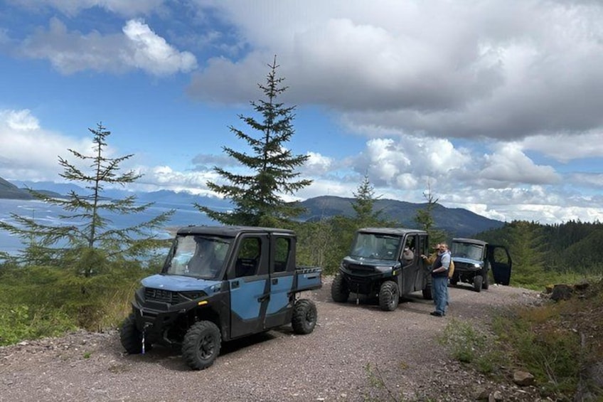 Two off-road vehicles parked on a gravel path with a lake and mountains in the background. 1