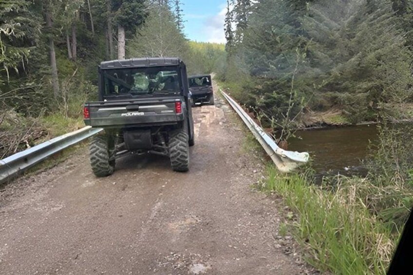A black off-road vehicle on a dirt path beside a narrow bridge over a stream, surrounded by dense forest. 3