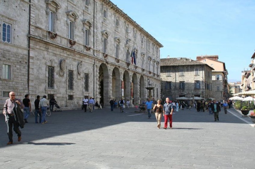 A historic stone building with arches, a paved square, and people walking. 4