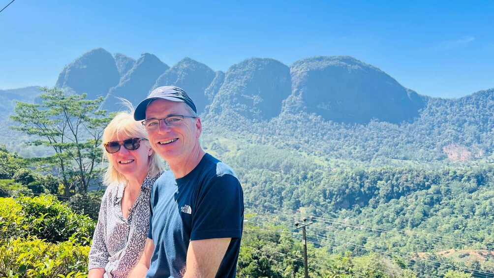 Two people standing in a mountainous area with dense forest and rocky outcrops in the background. 3