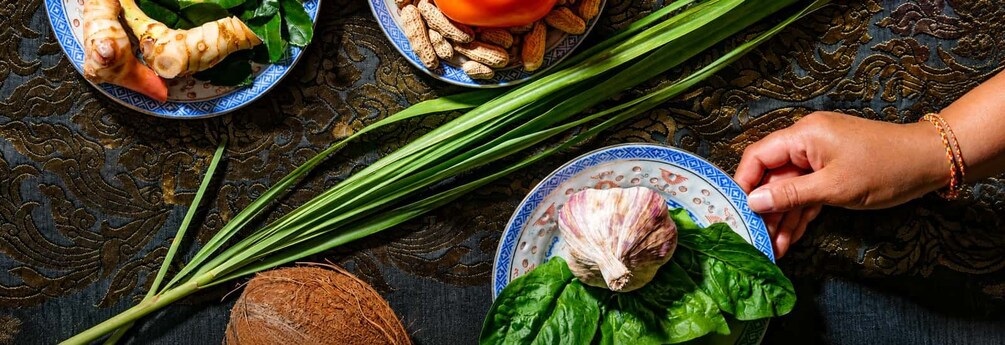 Une personne dispose un chou sur une assiette, à côté d’une feuille de laitue, le tout étant entouré d’oignons verts et d’une noix de coco, sur une nappe à motifs. 1