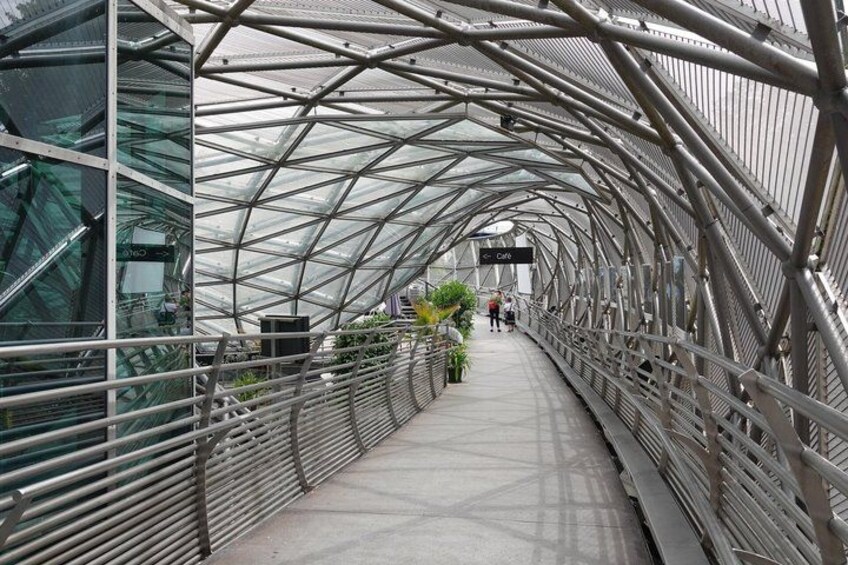 A pedestrian bridge with a metal framework and glass panels, featuring a cafe area in the distance. 4