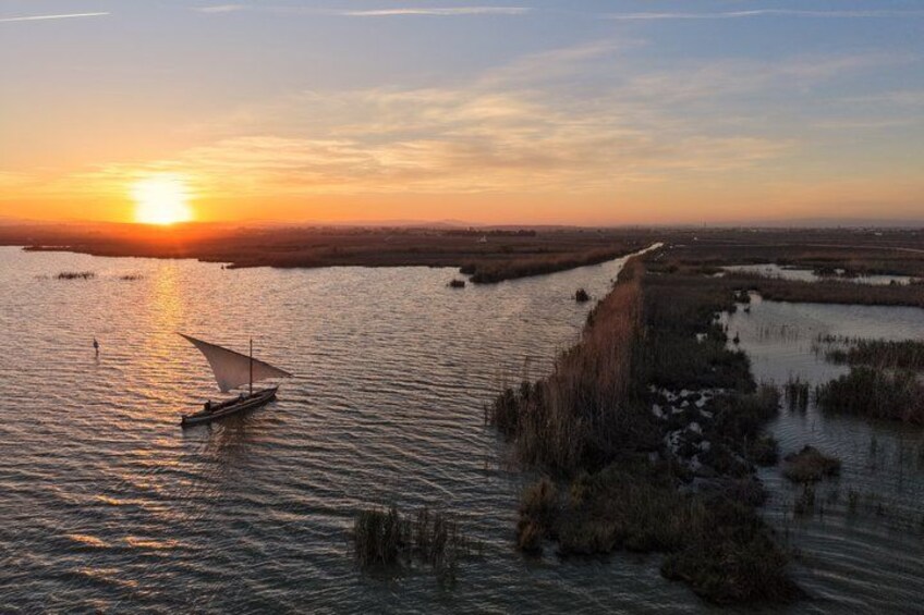A sunset over a water body with a sailboat and marshland. 2
