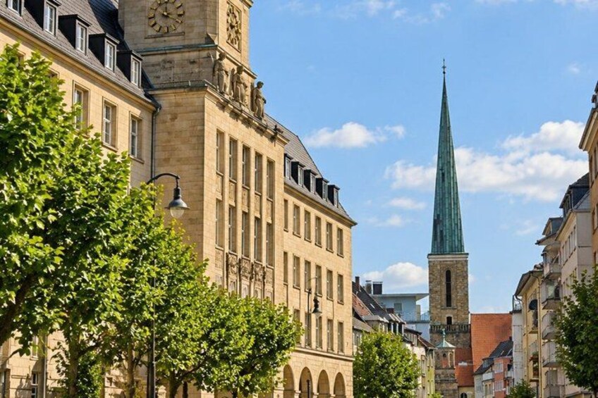 A historic building with a clock tower, a church spire, and trees lining the street. 4