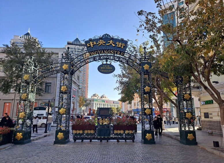 A street entrance with ornate black metal gates and golden decorations, leading to a cobblestone street lined with trees and buildings. 3