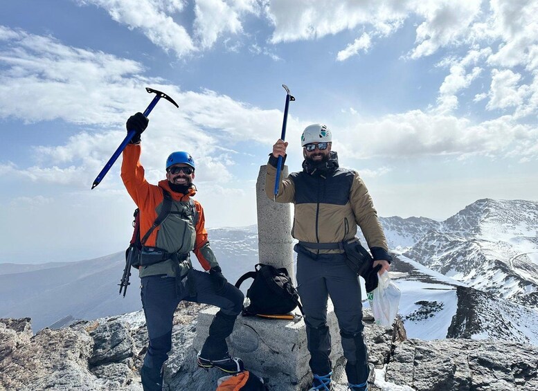 Two people standing on a rocky mountain peak with snow-covered terrain in the background. 1