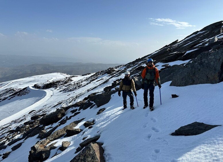 Two hikers on a snowy mountain trail with rocky terrain and distant mountains in the background. 5