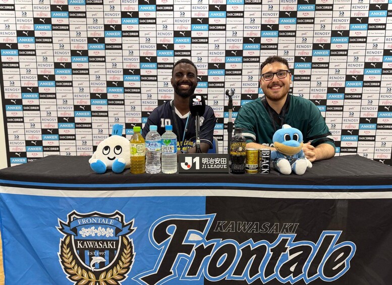 Two people at a table with sponsor logos, a plush toy, and a sign reading 'Kawasaki Frontale'. 4