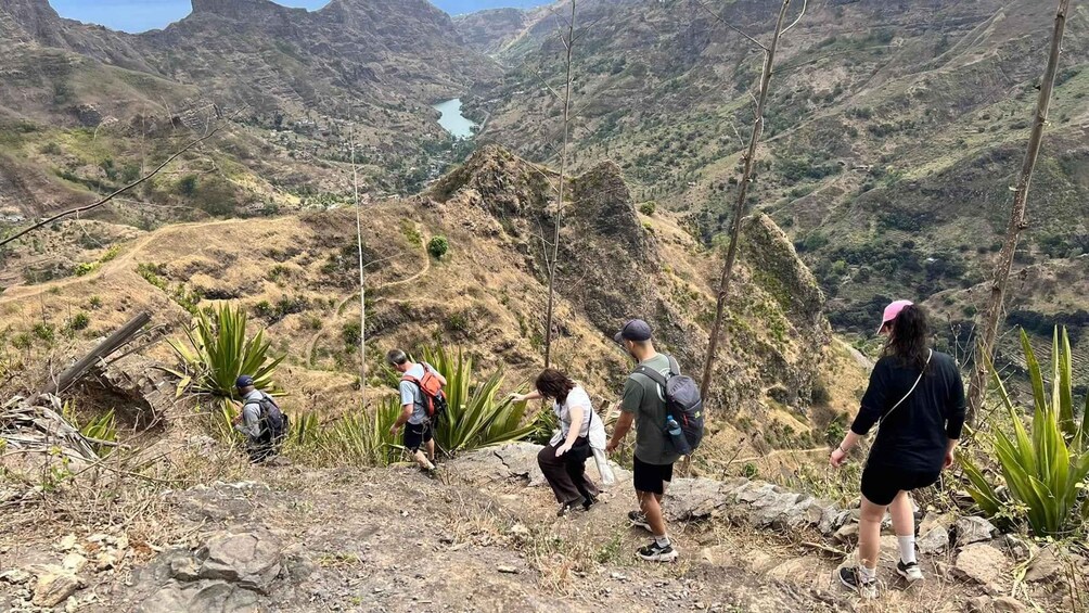 A group of hikers on a rocky trail with a mountainous landscape and a lake in the distance. 3