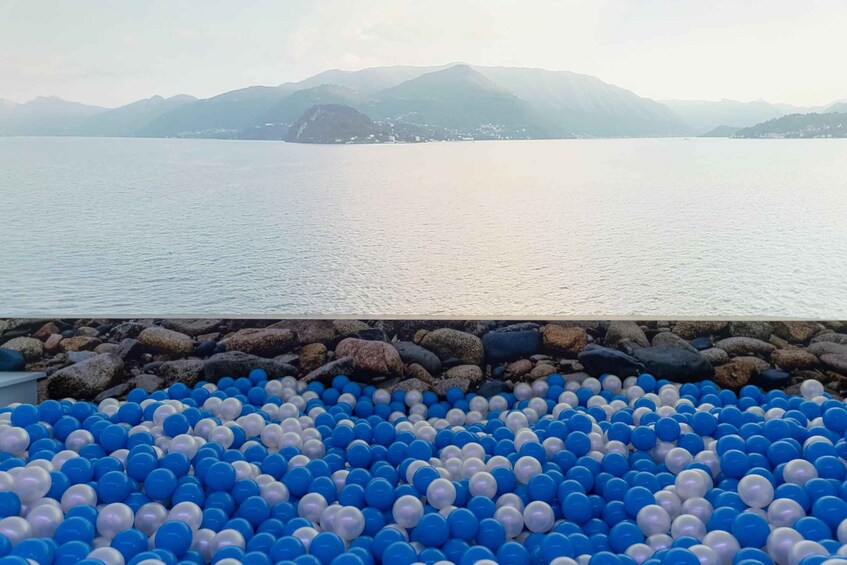 A body of water with mountains in the background and a beach with blue and white plastic balls. 1