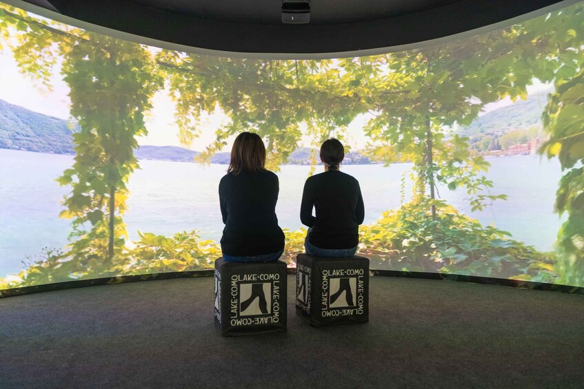 Two people sitting on stools facing a large screen displaying a scenic view of a lake surrounded by trees. 5