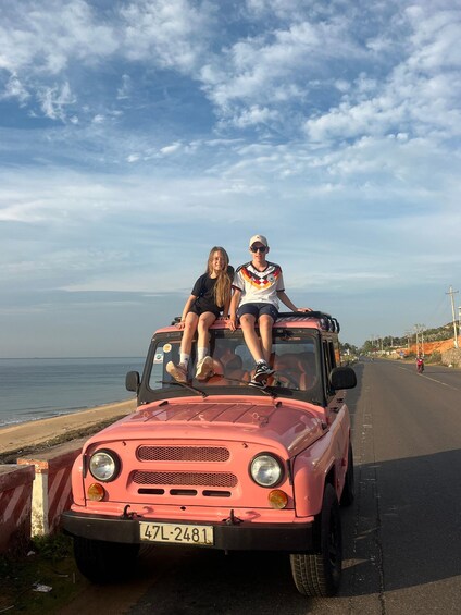 Une voiture ancienne rose, avec deux personnes assises sur le toit, est garée sur une route côtière, avec la mer en arrière-plan. 2
