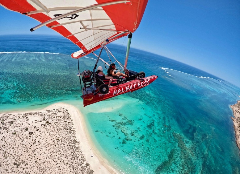 A hang glider with 'MALBAT ROSS' written on it flying over a turquoise sea and a sandy beach. 2