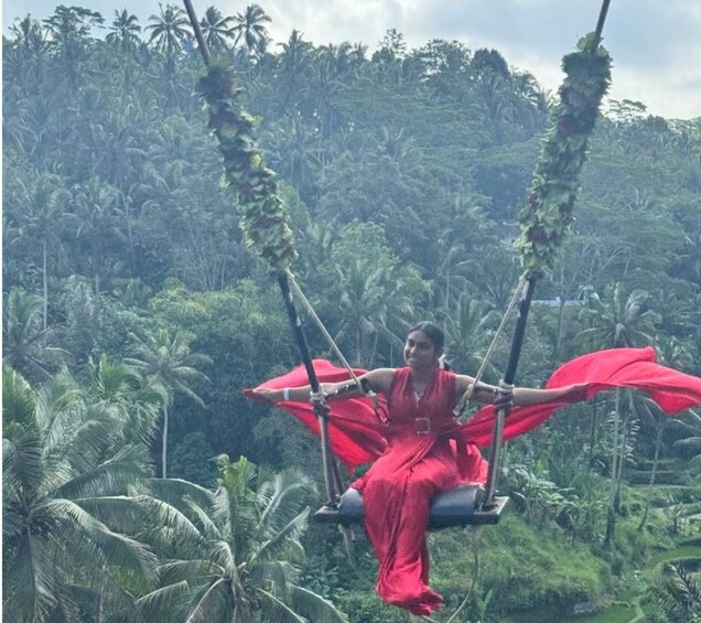 Une personne vêtue d’une robe rouge est assise sur une balançoire dont l’assise est également en tissu rouge, et elle est entourée d’une forêt tropicale dense. 2