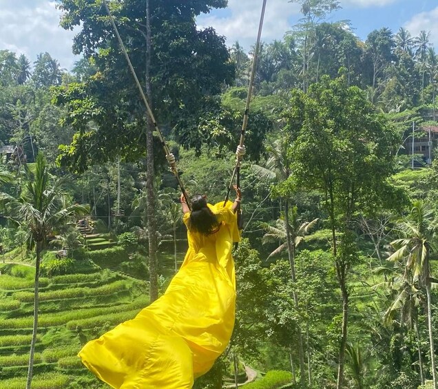 Une personne vêtue d’une robe jaune se balance sur une corde, entourée d’une végétation luxuriante et de champs en terrasses. 3