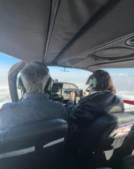 Two people inside an aircraft cockpit with a view of the sky and clouds outside. 1