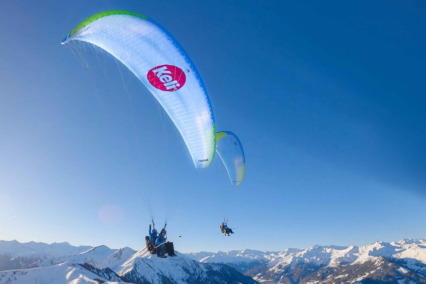 A paraglider with a white canopy and a logo, flying over a snowy mountain range. 2