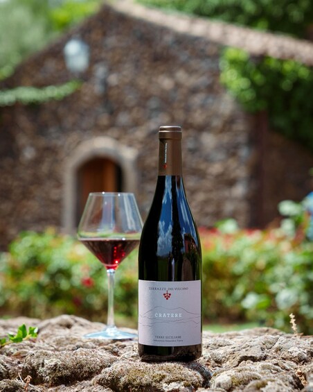 En flaska Terrazze del Vulcano Cratere-vin tillsammans med ett glas rött vin, placerat framför en rustik stenbyggnad med grönska i bakgrunden. 3