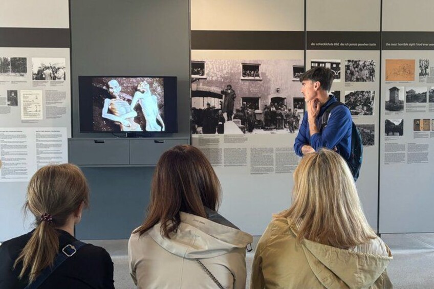 A museum exhibit with a television screen displaying a historical scene, informational panels, and visitors observing. 5