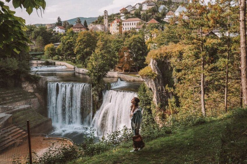A waterfall, a person standing on a grassy area, and houses in the background. 1