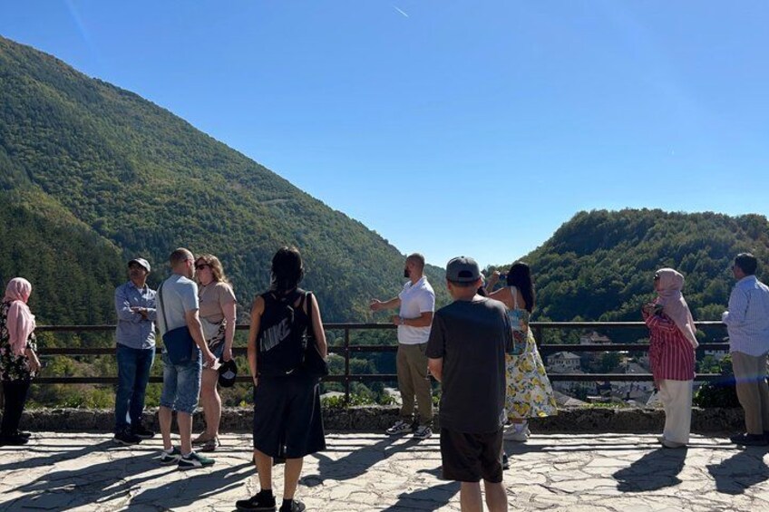 A group of people standing on a stone bridge overlooking a river and a forested hill. 2