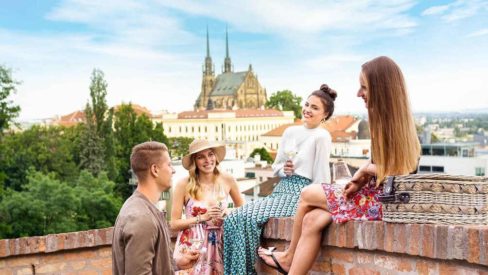 Eine Gruppe von Menschen sitzt auf einer Ziegelmauer und blickt auf eine Stadtlandschaft mit einem markanten Kirchturm. 1