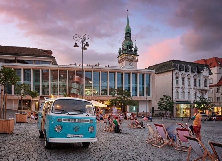 Ein blauer Van parkt auf einem Kopfsteinpflasterplatz, auf dem Stühle stehen. Im Hintergrund ist ein Kirchturm zu sehen, und ein modernes Gebäude mit großen Fenstern. 2