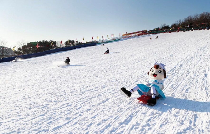 A snow-covered slope with people skiing and snowboarding, a mascot sitting on the ground, and flags lining the top. 5