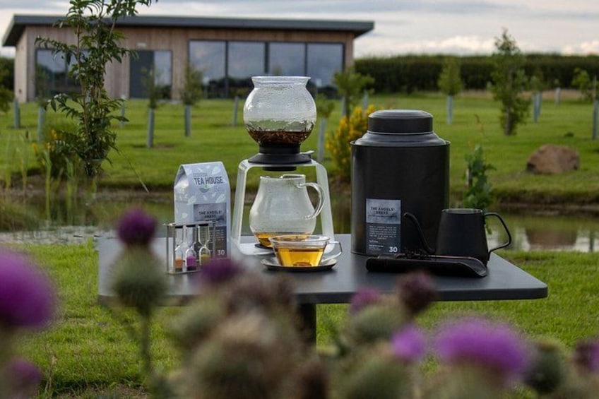 A table with a tea bag, a glass pitcher, a thermos, and a mug in a garden setting. 4