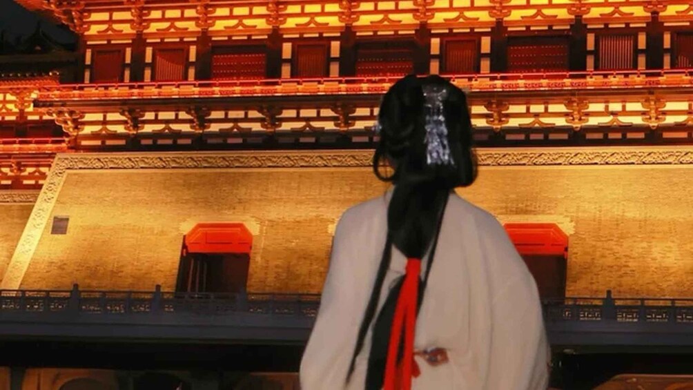 A person with a long black and white headpiece standing in front of a traditional building with red accents. 2