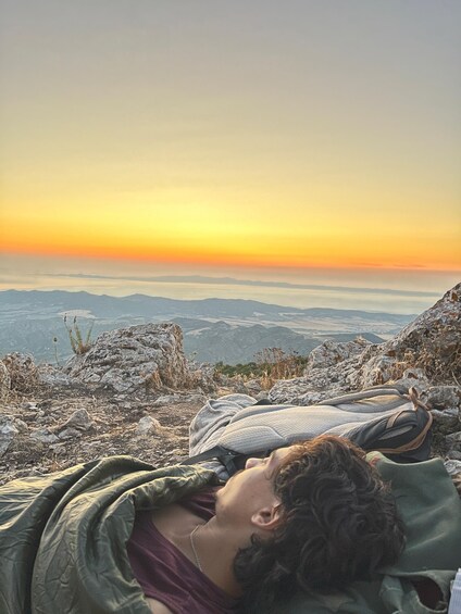 Une personne allongée sur une couverture, sur une montagne rocheuse, d’où l’on a une vue sur une vallée au coucher du soleil. 1