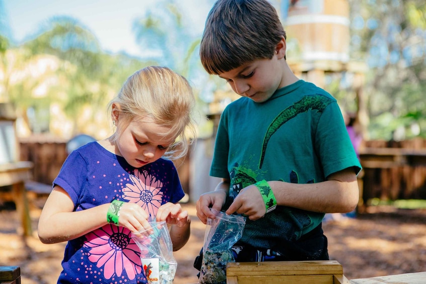 Two children outdoors, one wearing a blue shirt with a floral design and the other in a green shirt, handling plastic bags with items. 5