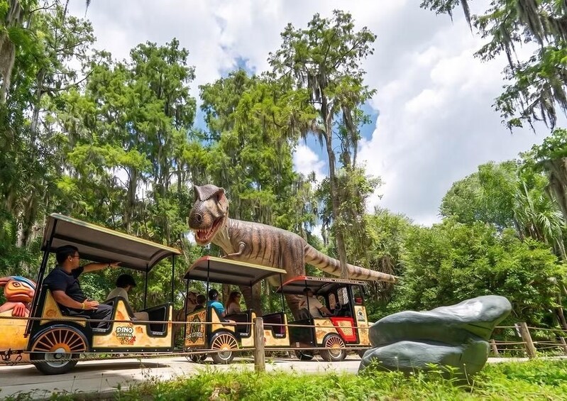 A dinosaur-themed ride with passengers, surrounded by lush greenery and trees. 1