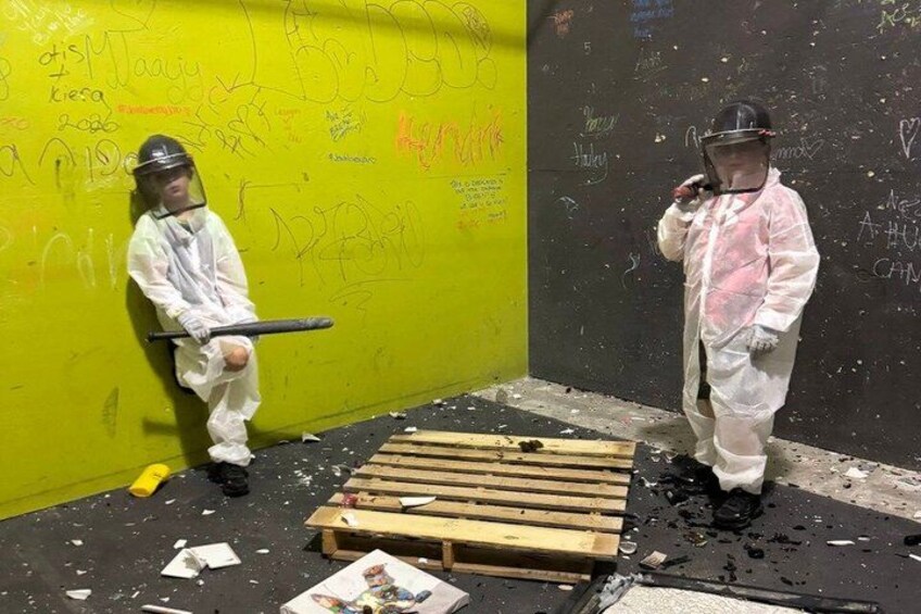 Two people in protective suits, one holding a bat, standing in a room with graffiti-covered walls and scattered debris. 3