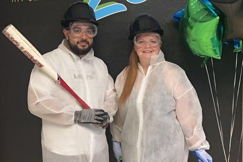 Two people in protective gear, one holding a cricket bat, standing in front of a wall with a logo and balloons. 5