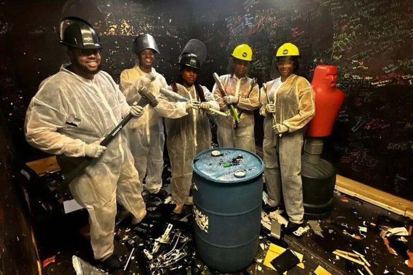 A group of people in protective suits and helmets, holding tools, in a room with a large blue barrel and scattered debris. 1