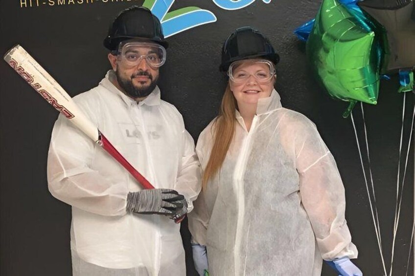 Two people in protective gear holding a cricket bat, with a logo and balloons in the background. 4